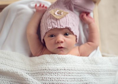 Newborn baby lying in toy bed with hands over her head and eyes wide open wearing pink cap | san diego newborn photography | www.babiesandbeauties.com