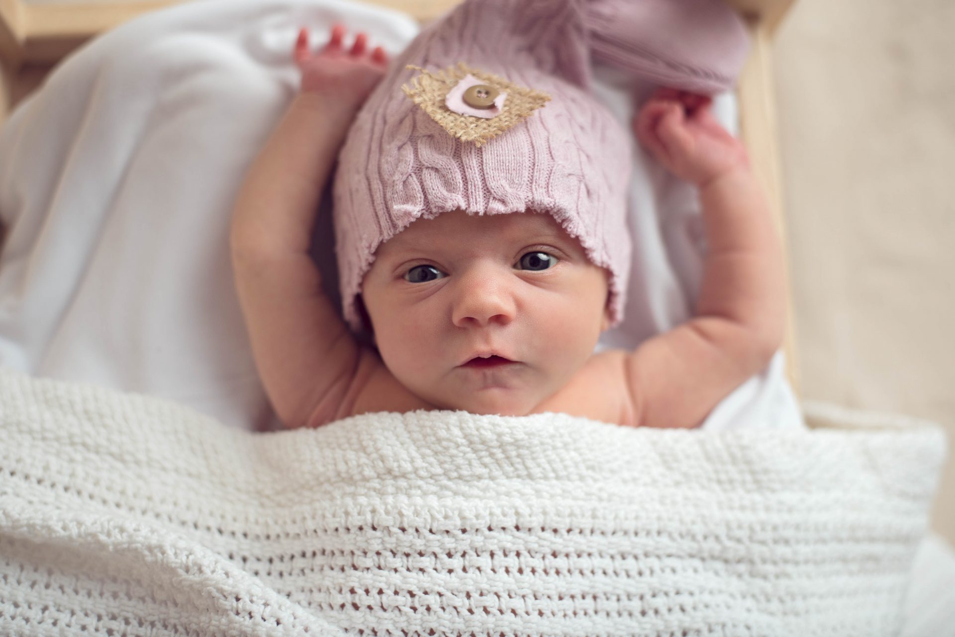 Newborn baby lying in toy bed with hands over her head and eyes wide open wearing pink cap | san diego newborn photography | www.babiesandbeauties.com