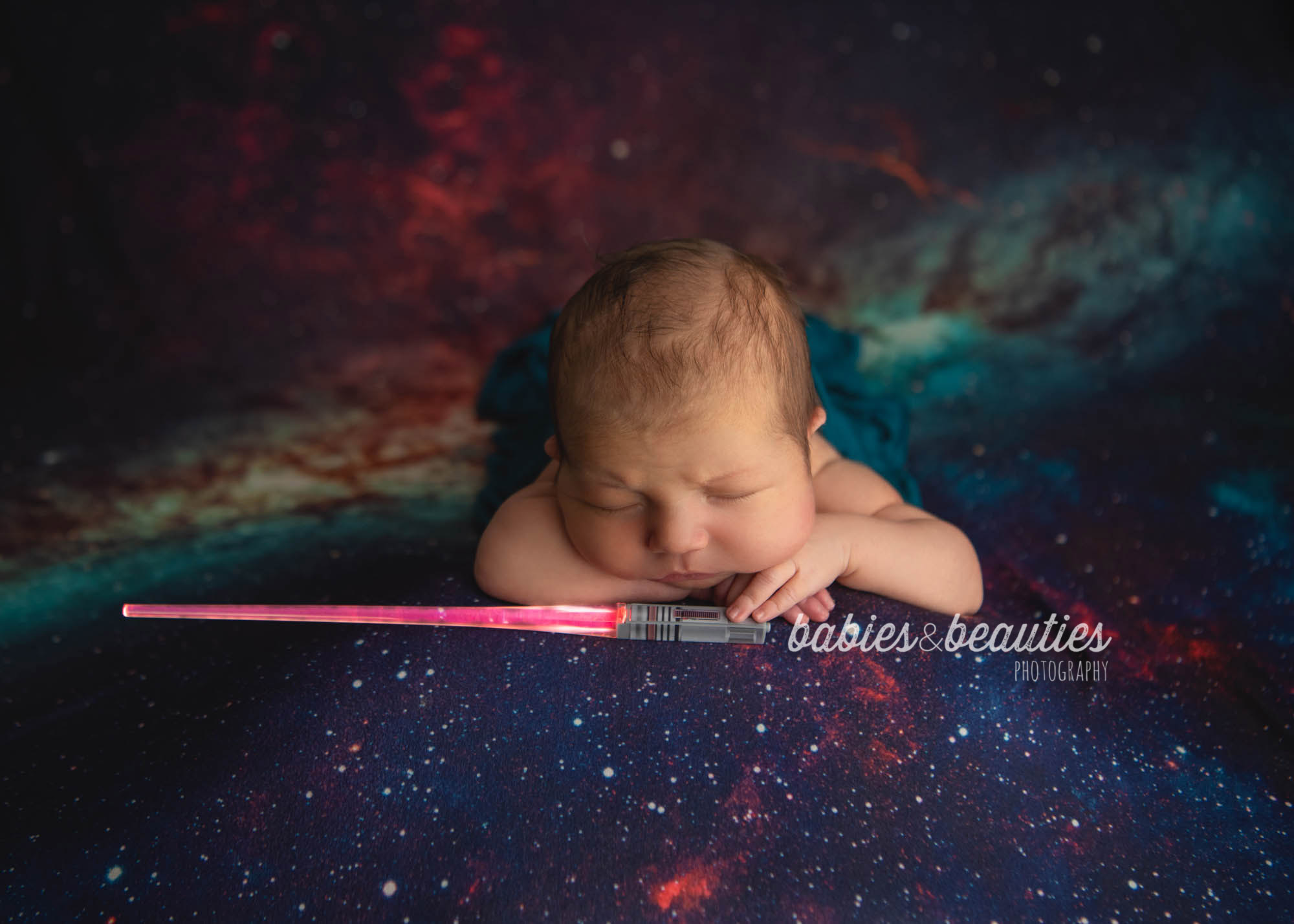 Newborn photography of little boy on galaxy background with tiny light saber. Book your San Diego newborn photography session today! www.babiesandbeauties.com